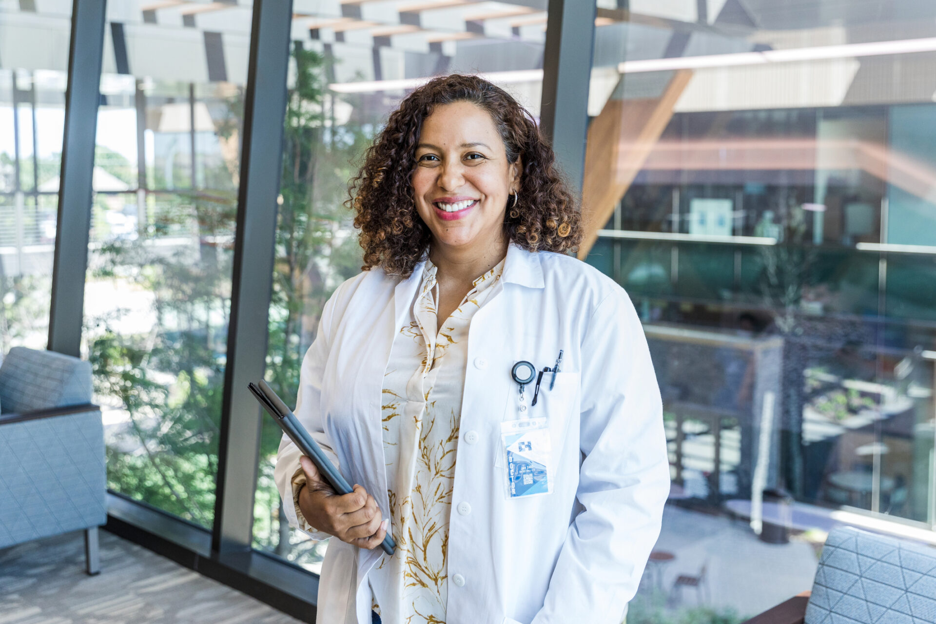A smiling woman with curly hair wearing a white lab coat and holding a folder stands in a bright, modern office with large windows—reflecting the professional environment of nursing jobs at Covenant Health.