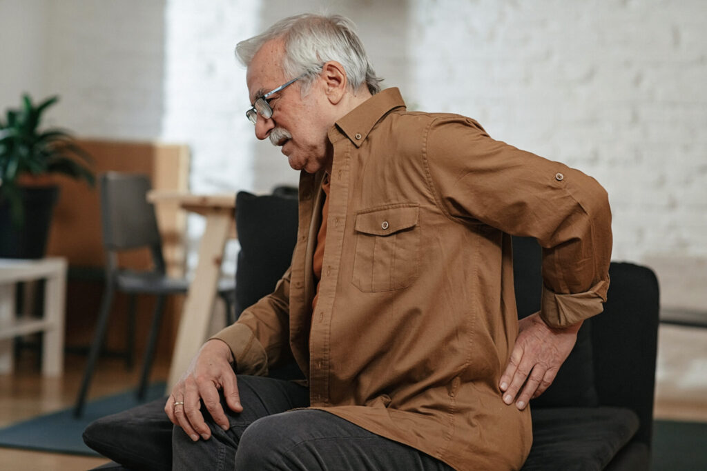 An older man with gray hair and glasses sits on a couch, holding his lower back with one hand and appearing to be in pain. The background shows a bright living room with a plant and chairs.