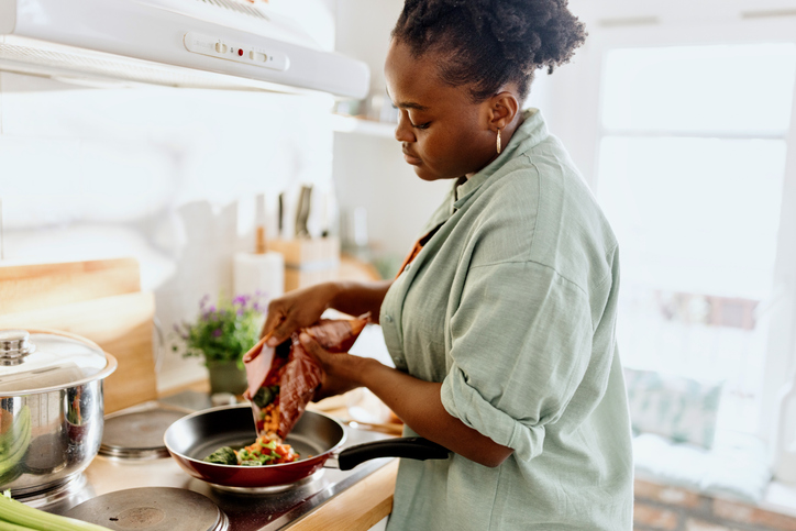 A woman in a light green shirt is cooking in a kitchen, adding ingredients from a package into a frying pan on the stove. Sunlight streams in through a window in the background.
