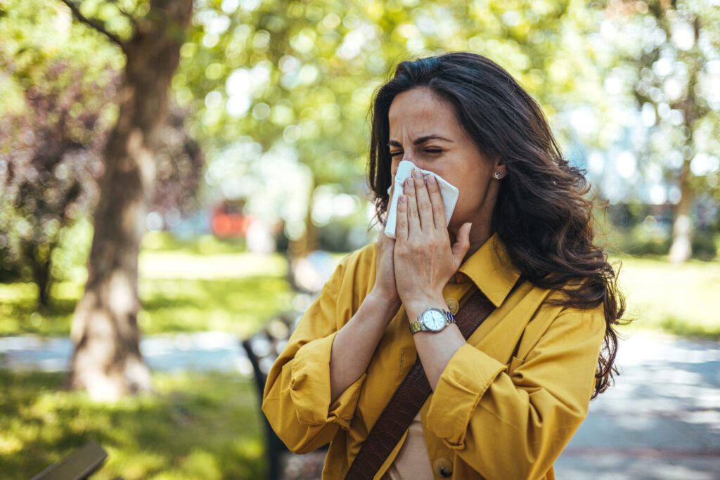 A woman standing outdoors in a park sneezes into a tissue, wearing a yellow jacket. Trees and sunlight are visible in the background, suggesting a spring or summer day.