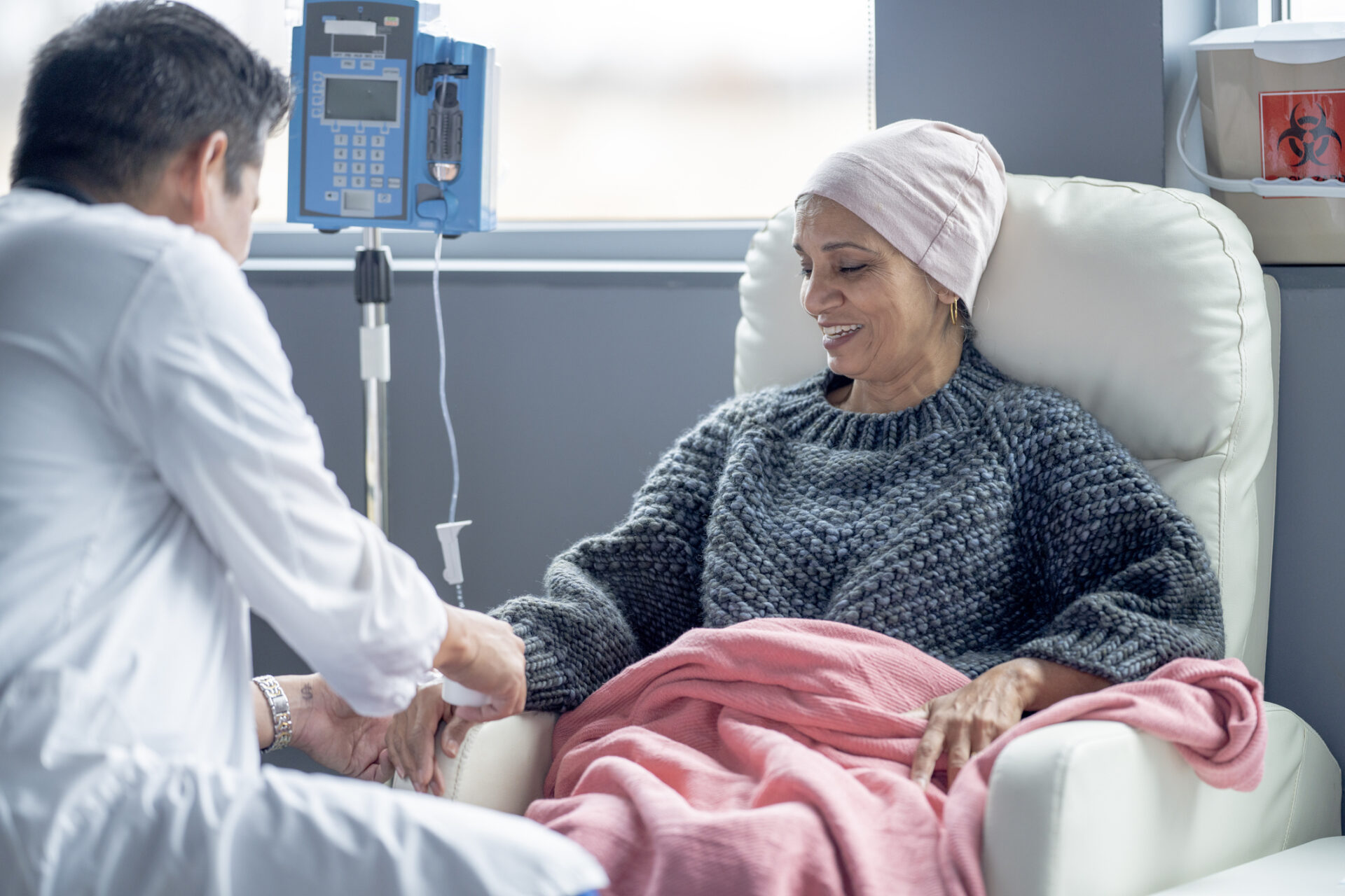 A woman wearing a headscarf sits in a hospital chair, covered with a pink blanket, smiling while receiving IV treatment from a healthcare professional.