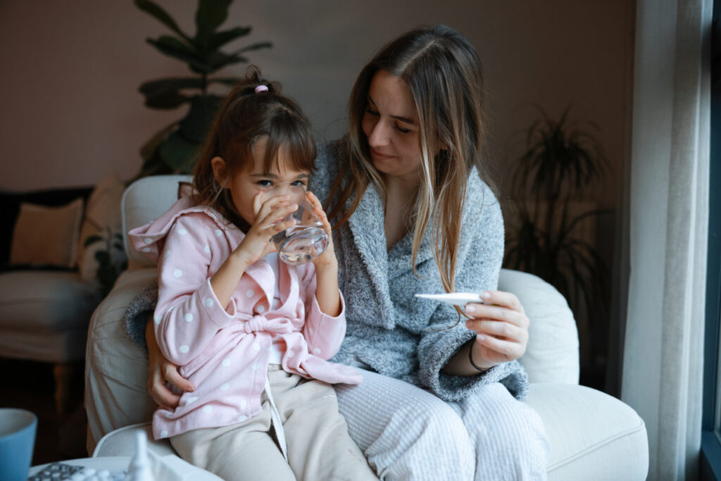 A woman in pajamas holds a thermometer and sits on a couch with a young girl in a pink robe, who is drinking water. They appear to be at home, caring for the girl as she recovers from a respiratory illness amid plants and soft lighting.