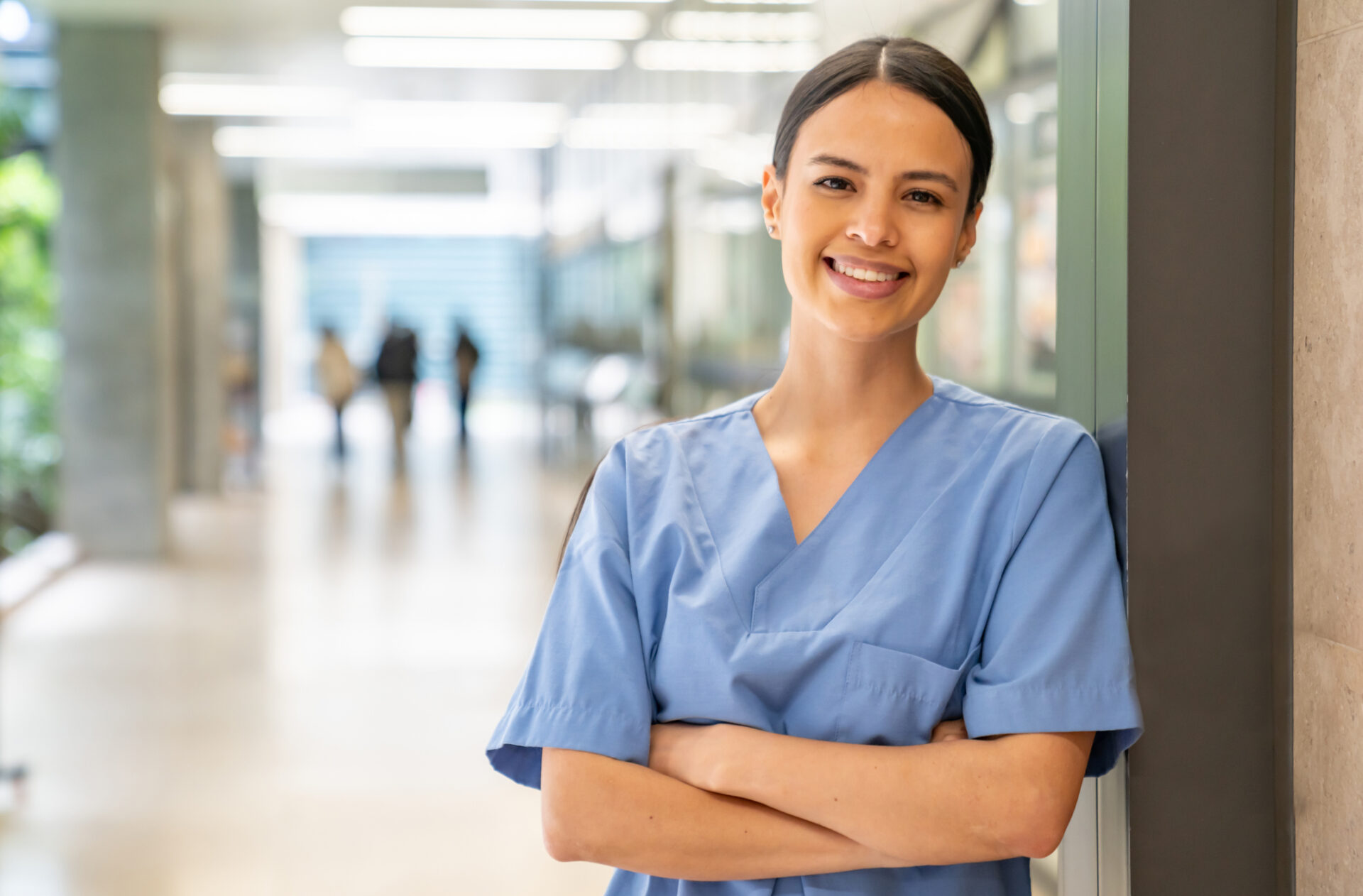 A smiling primary care healthcare worker in blue scrubs stands with folded arms in a bright hospital corridor, with blurred people walking in the background.