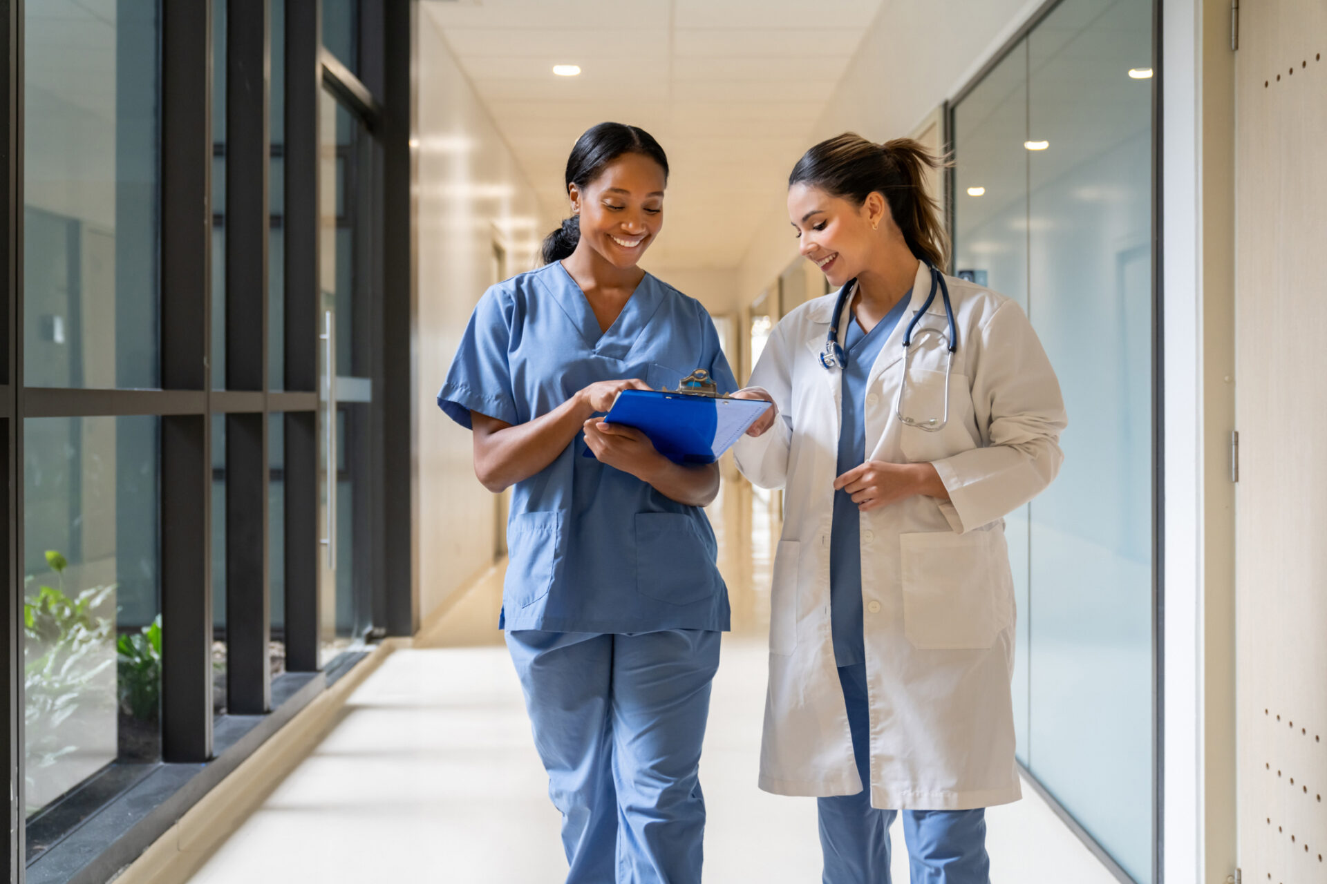 Two female healthcare professionals walk down a hallway in a primary care clinic. One wears blue scrubs and holds a clipboard, while the other wears a white lab coat and stethoscope. Both are smiling and appear to be discussing patient notes.