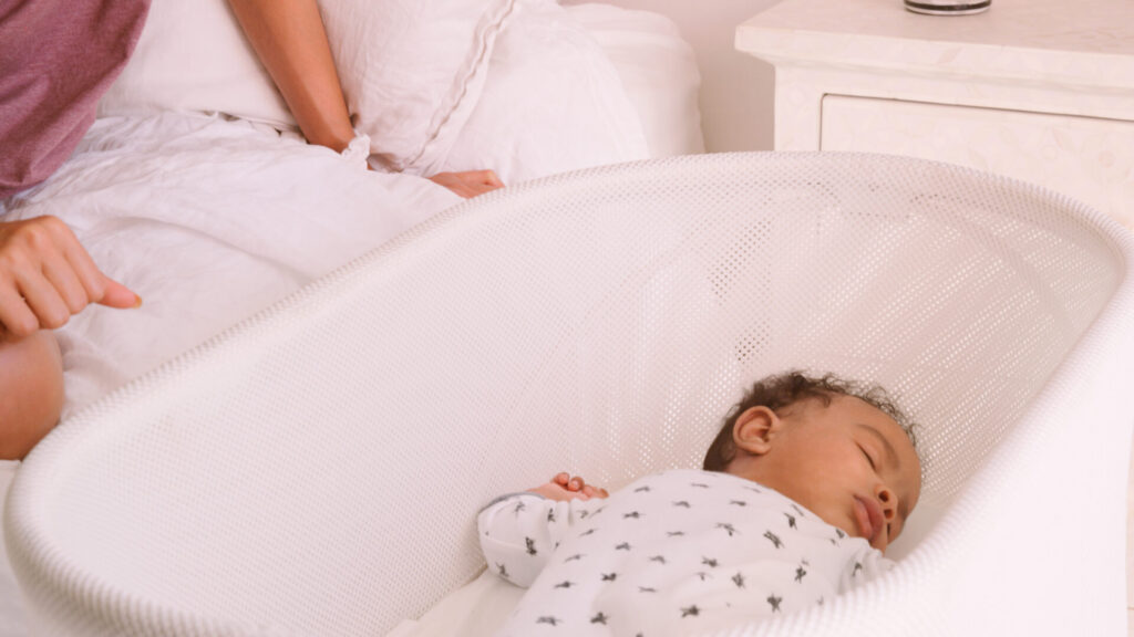 A baby sleeps peacefully in a white bassinet while an adult sits on a nearby bed, partially visible. The scene is calm and softly lit, with white bedding and a nightstand in the background.