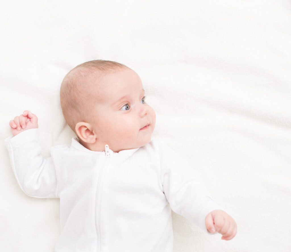 A baby with light skin and blue eyes lies on a soft white blanket, wearing a white outfit, looking to the side with one arm raised. The background is bright and minimal.