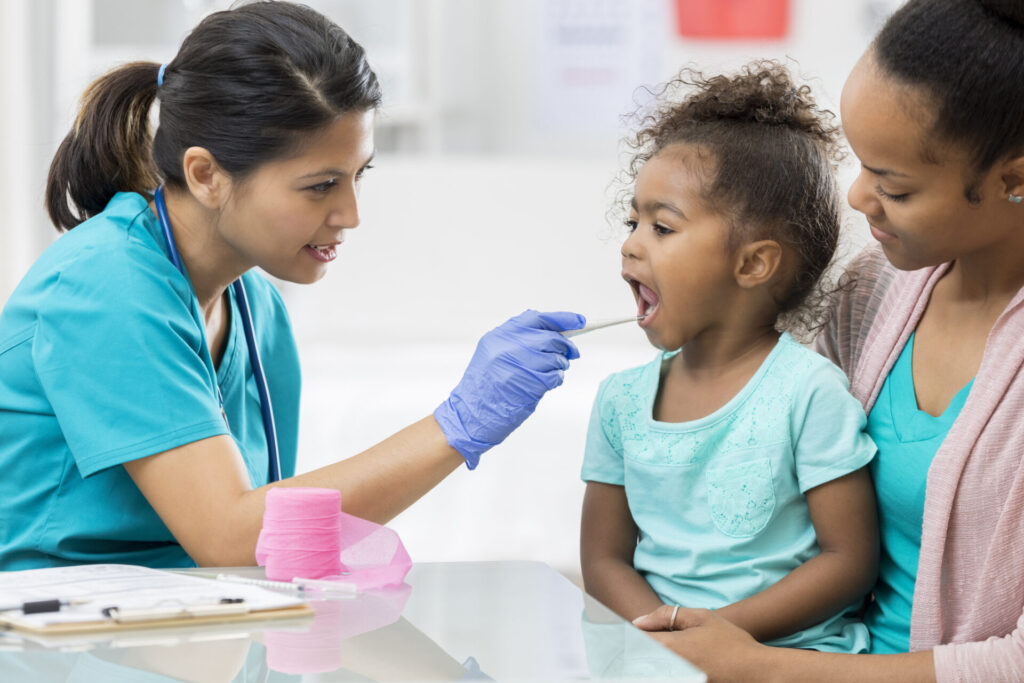 A female healthcare worker in scrubs examines a young girl&rsquo;s sore throat with a tongue depressor while the child, sitting on a woman&rsquo;s lap, is comforted; both wear teal shirts in a medical office.