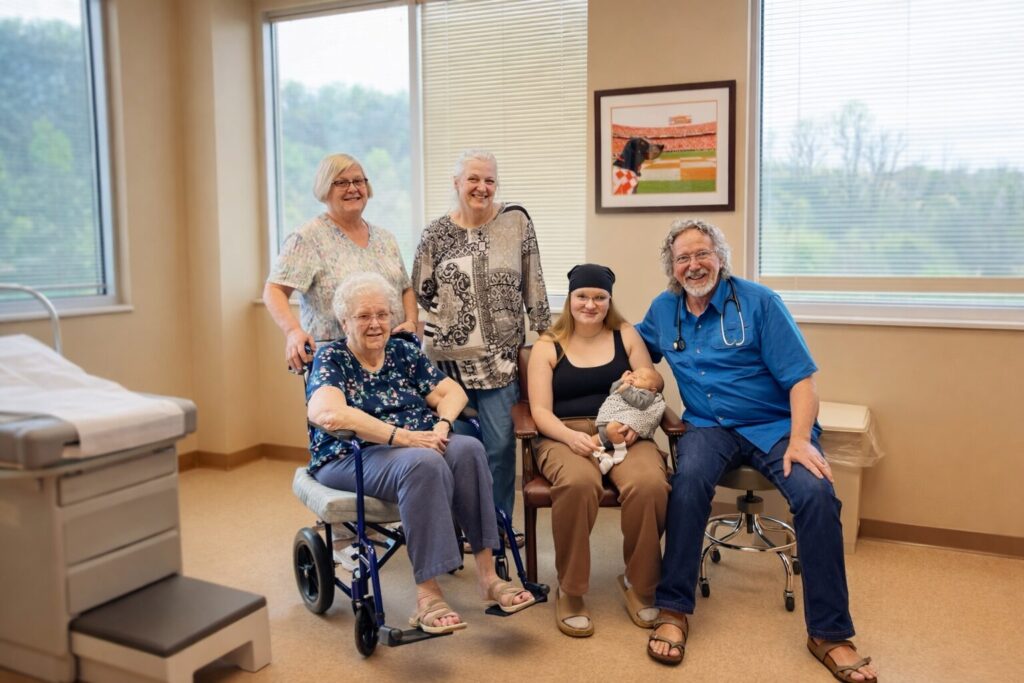 A group of five adults and one baby pose and smile in a medical office. One woman sits in a wheelchair, another holds a baby, and a man with a stethoscope sits beside them. Large windows and framed art are in the background.