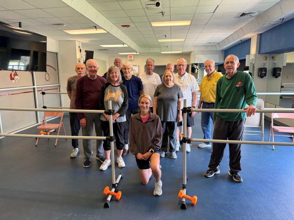 A group of eleven older adults and one younger woman kneel and stand together, smiling inside a brightly lit physical therapy or rehabilitation room with parallel bars and chairs.