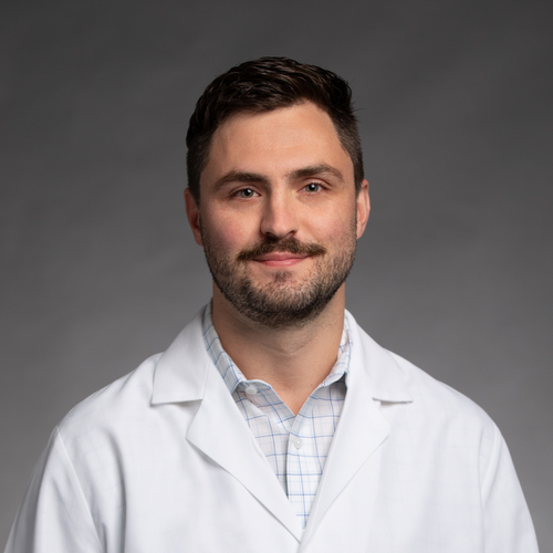 A man with short brown hair and a beard, wearing a white lab coat over a plaid shirt, stands in front of a plain gray background, smiling slightly at the camera.