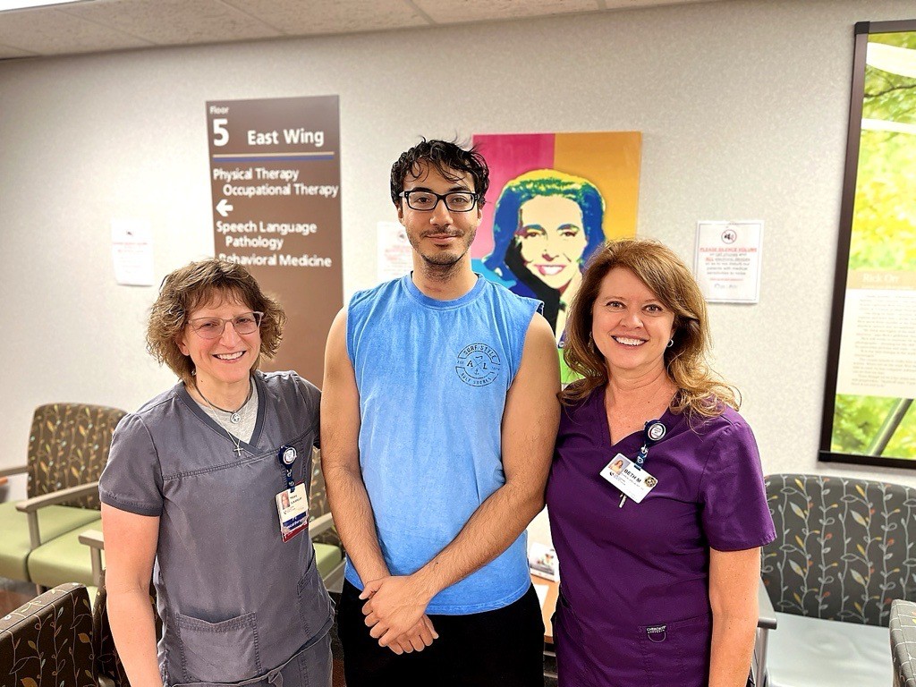 Three people, two women in medical scrubs and a man in a sleeveless blue shirt, stand smiling in a waiting area with a colorful portrait and healthcare signs in the background.