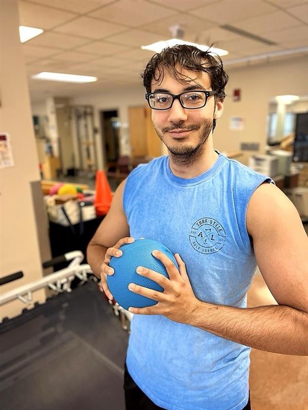 A young man wearing glasses and a blue sleeveless shirt stands indoors holding a blue textured ball, smiling at the camera. Gym equipment and exercise cones are visible in the background.