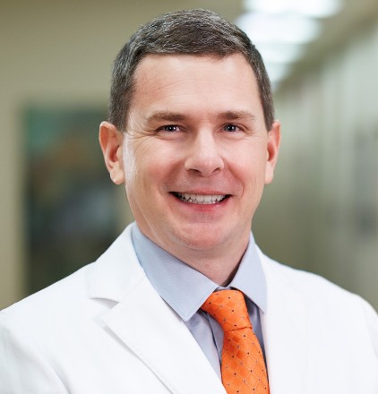 A smiling man in a white lab coat and an orange tie stands in a well-lit hallway, suggesting a professional or medical setting.