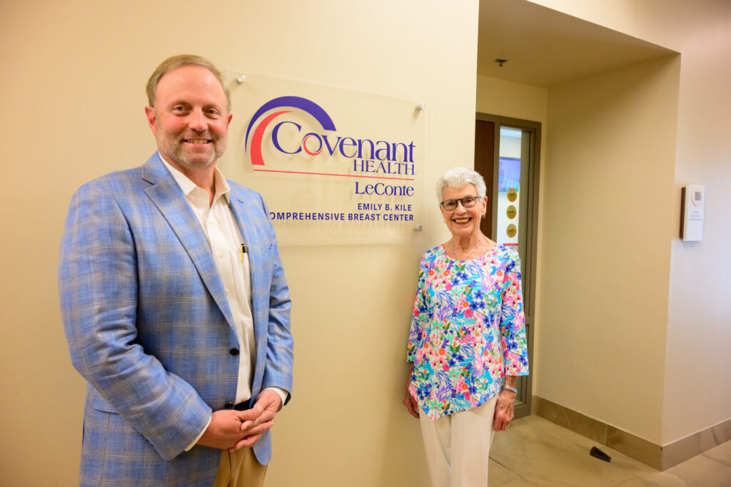 A man in a light blue plaid jacket and a woman in a colorful floral top stand smiling beside a Breast Center sign that reads Covenant Health LeConte Comprehensive Breast Center in a hallway.