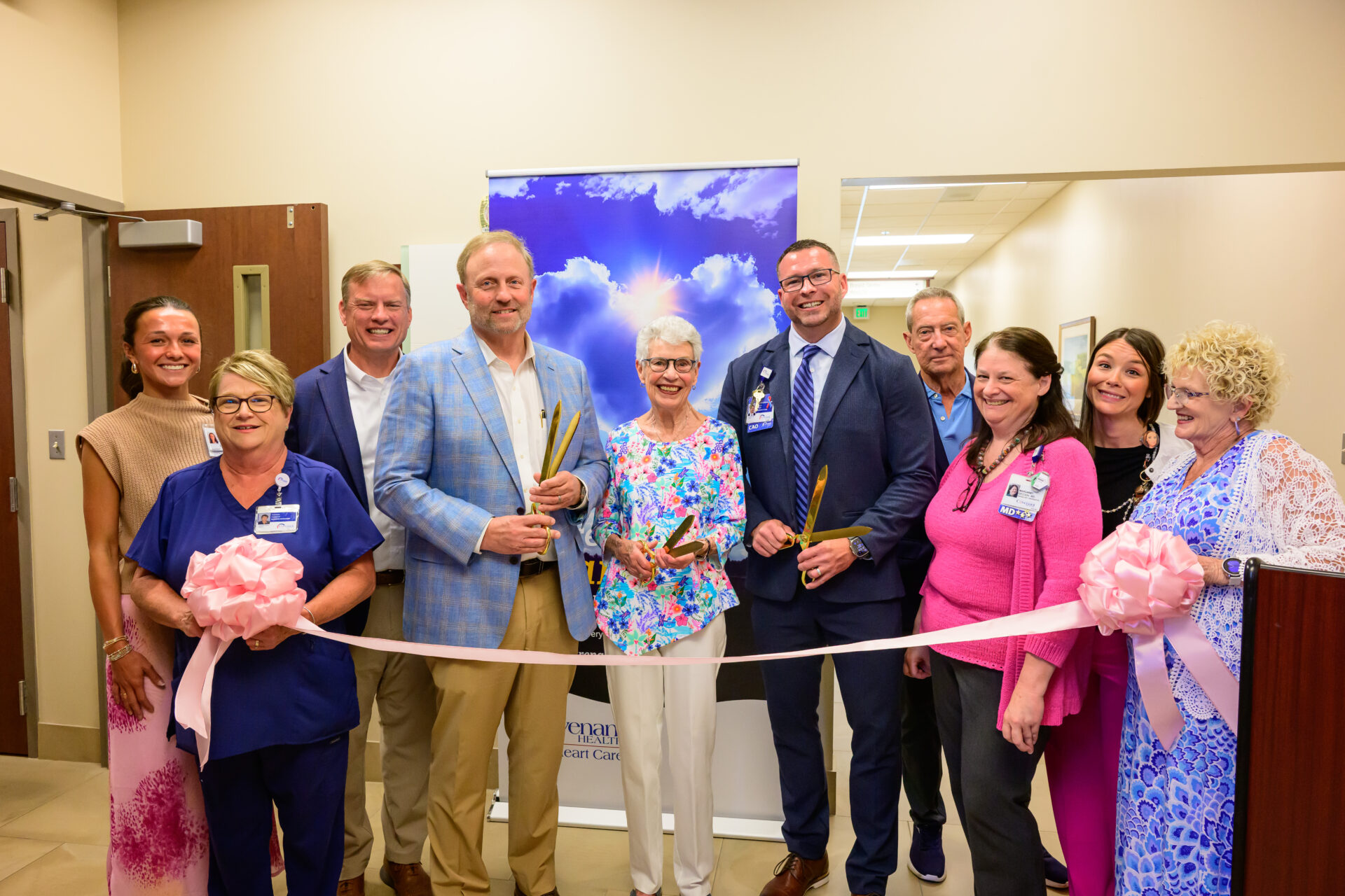 A group of people smile at a ribbon-cutting ceremony indoors. Three people in the center hold oversized scissors, while others stand around them. A pink ribbon and bows are in front, with a blue sky banner in back.