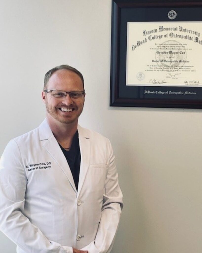 General surgeon Wayne Cox, in a white lab coat, stands smiling next to a wall-mounted diploma from Lincoln Memorial University, DeBusk College of Osteopathic Medicine.