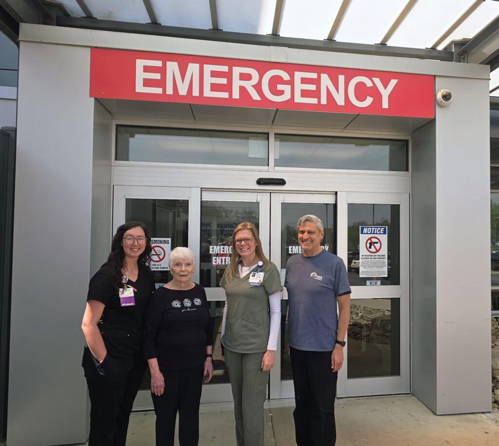 Four people stand smiling outside a hospital emergency entrance under a red “EMERGENCY” sign. Two women wear scrubs, one wears a black shirt, and one person, with gray hair, wears a dark polo shirt.