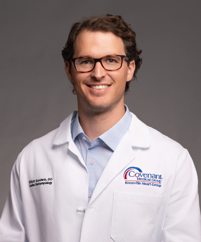 A man with short brown hair and glasses smiles at the camera. He is wearing a white lab coat with the Covenant Medical Group Knoxville Heart Group logo and an embroidered name tag over a light blue collared shirt, standing against a gray background.