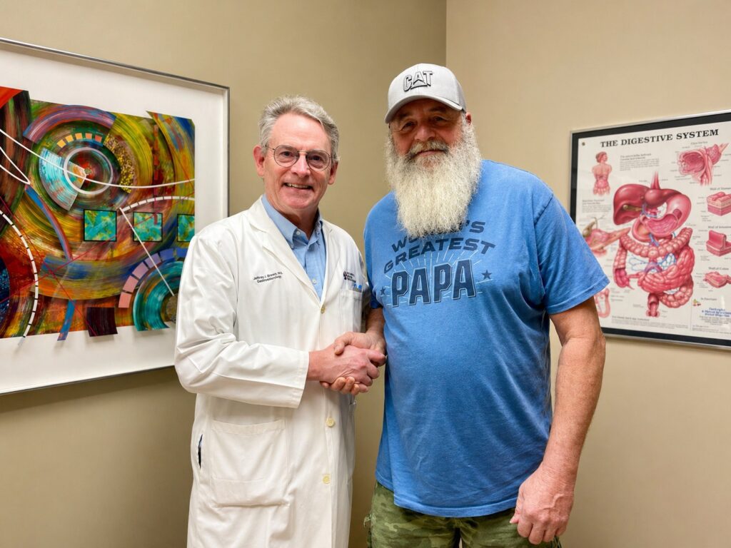 A doctor in a white coat shakes hands with a bearded man wearing a blue Worlds Greatest Papa T-shirt and a light gray hat, standing in a medical office with colorful and anatomical posters on the walls.