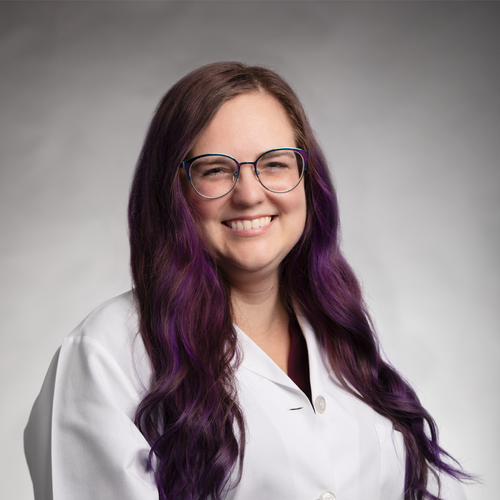 A woman with long, wavy purple hair and glasses smiles while wearing a white lab coat, standing against a plain light gray background.
