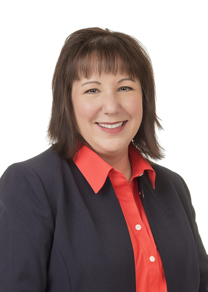 A woman with straight brown hair wearing a navy blazer over a bright coral blouse, smiling at the camera against a plain white background.