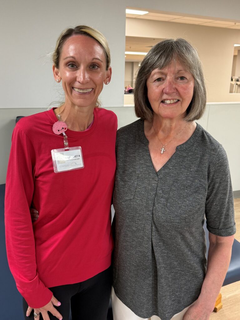 Two women standing close together and smiling indoors. The woman on the left wears a red long-sleeve shirt and a name badge, while the woman on the right wears a gray short-sleeve shirt and a cross necklace.