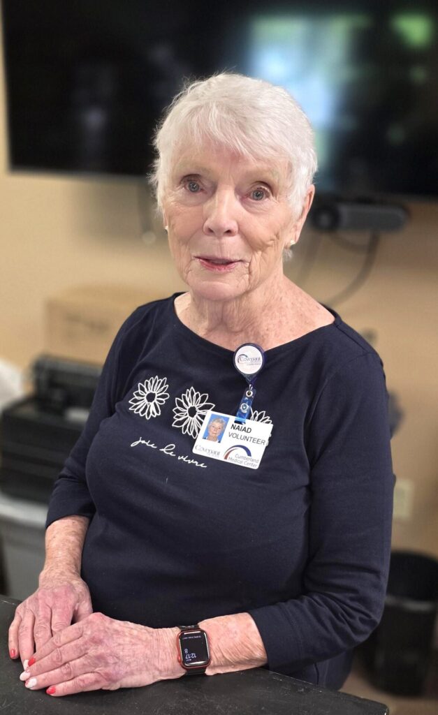 An older woman with short white hair, wearing a navy shirt and a volunteer badge, stands indoors. She has a smartwatch on her wrist and is smiling softly at the camera, recalling her recent visit to the emergency room for a tick bite. The background is blurred.