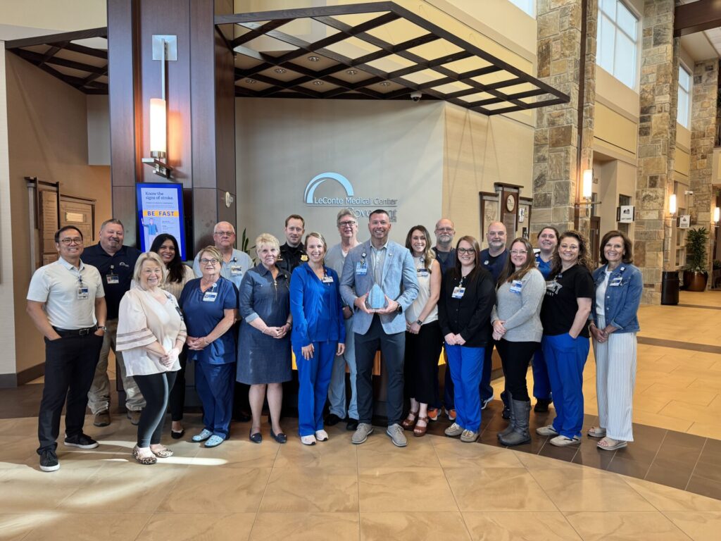 A group of hospital staff and professionals pose together in the lobby of a medical center, smiling at the camera. The group stands in front of a sign that reads LeConte Medical Center.