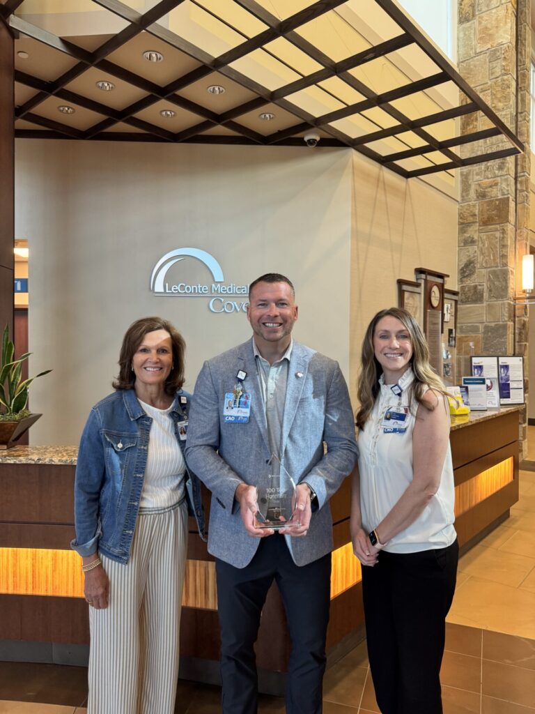Three people stand smiling in a lobby area with a sign reading LeConte Medical behind them. The person in the center holds a glass award. The group appears professionally dressed.