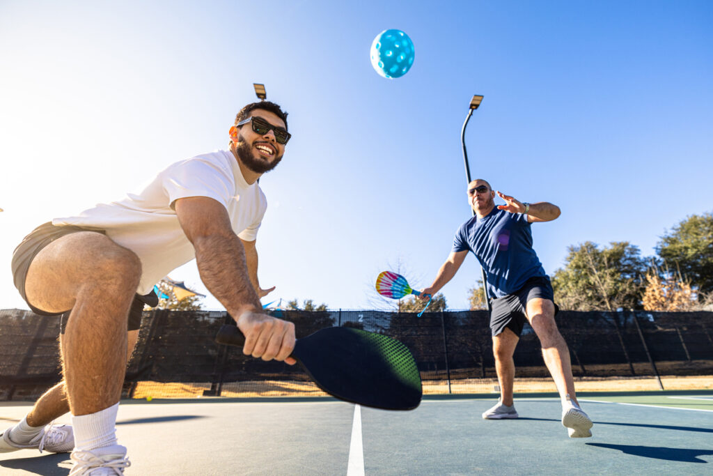 Two men play pickleball outdoors on a sunny day. One is lunging forward to hit a blue ball with his paddle while the other stands poised behind him, both smiling and wearing sunglasses. Trees and lights are visible in the background.
