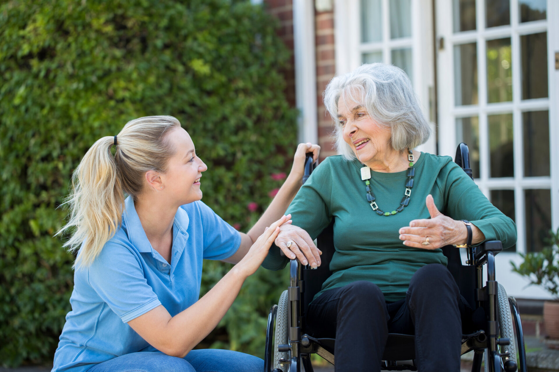 A young woman kneels beside an older woman in a wheelchair outdoors. They smile and engage in conversation in front of a house with greenery and glass doors in the background.