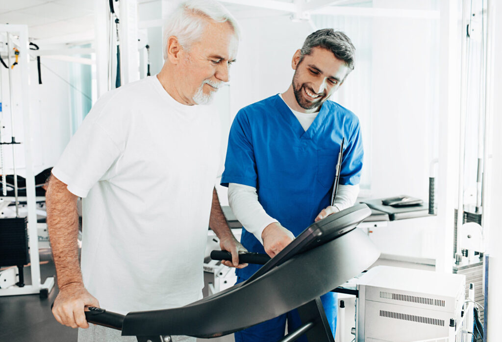 An older man walks on a treadmill while a healthcare professional in blue scrubs stands beside him, smiling and adjusting the machine in a bright, modern medical facility.