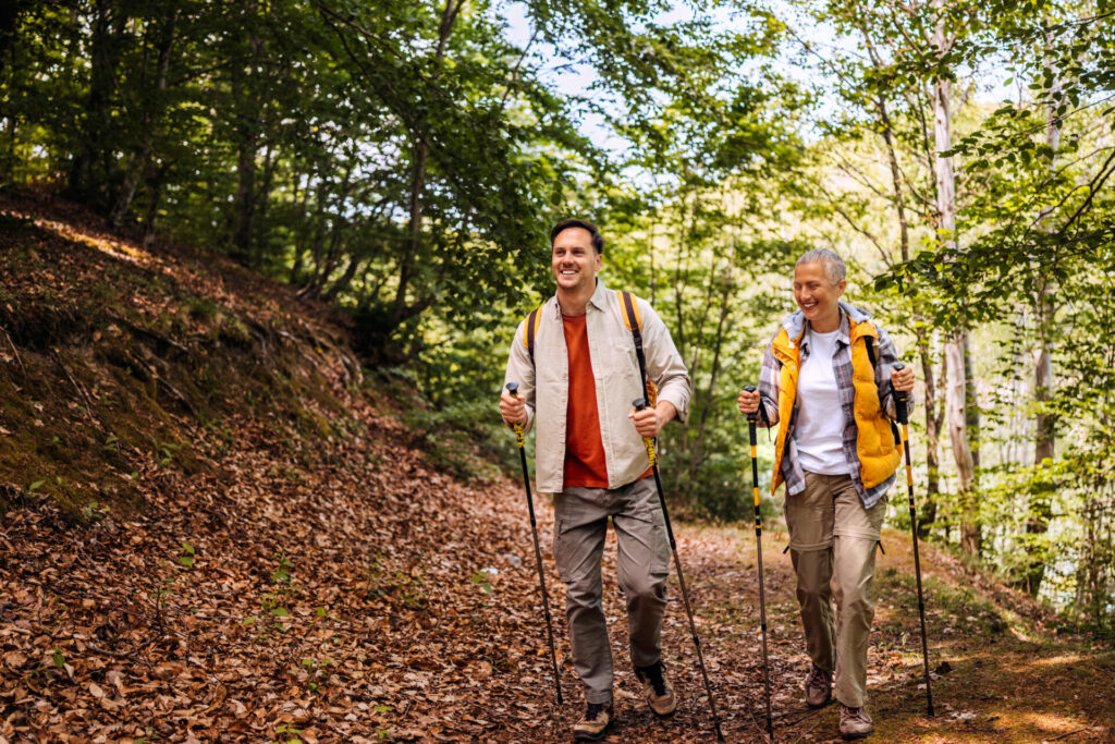 Two people with backpacks walk together on a forest trail, using trekking poles. They are surrounded by tall trees and fallen leaves, smiling and enjoying the outdoor hike in the sunlight.