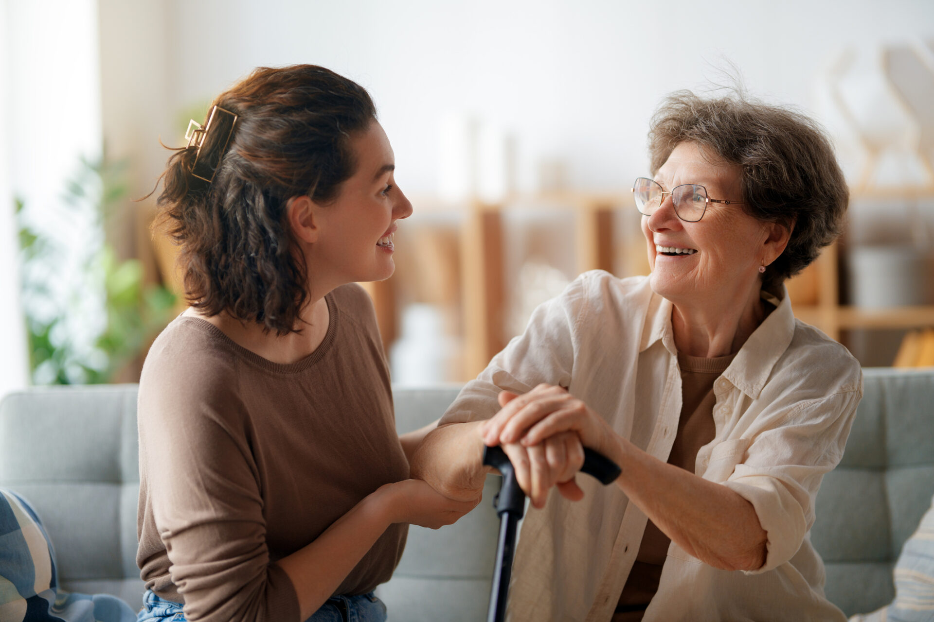 A young woman smiles and holds hands with an older woman using a cane. They are sitting together on a couch, sharing a joyful and warm moment in a bright, cozy living room.