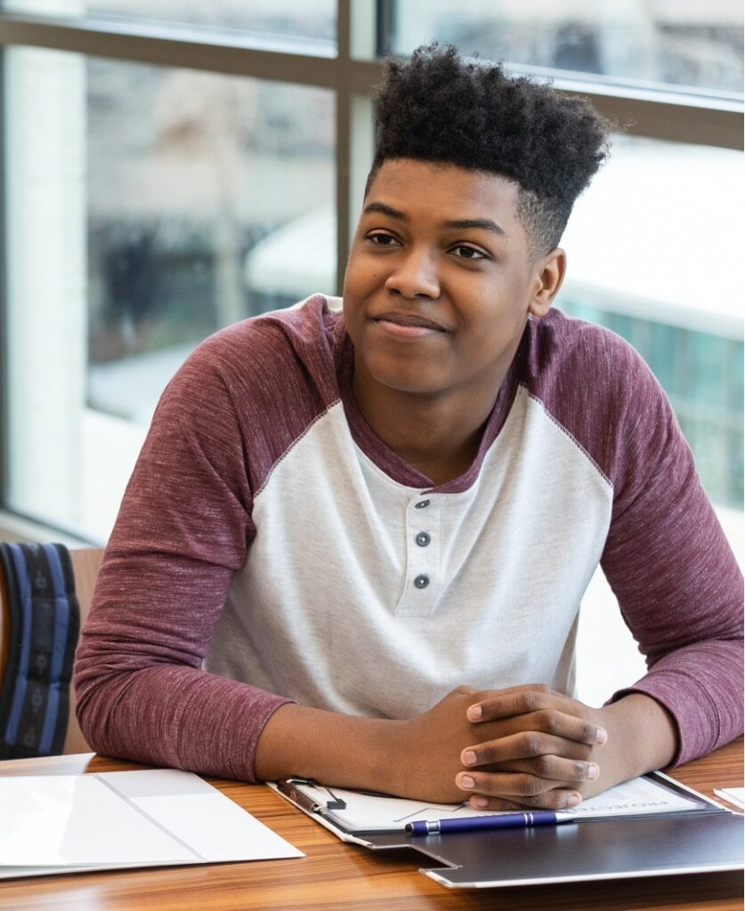 A student wearing a white and maroon long-sleeve shirt sits at a desk with papers, a clipboard, and a pen, smiling and looking slightly to the side in a bright room with large windows during parent information night at PCSA.