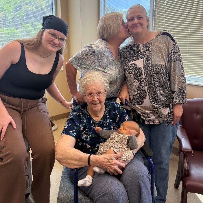 Four generations of women pose together indoors. An elderly woman sits in a wheelchair holding a baby, while three adult women stand behind her, one kissing another on the cheek. Large windows show greenery outside.