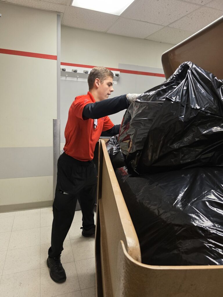 A person wearing a red shirt and black pants is pushing large black trash bags in a brown cart inside a room with tiled floor and beige walls.