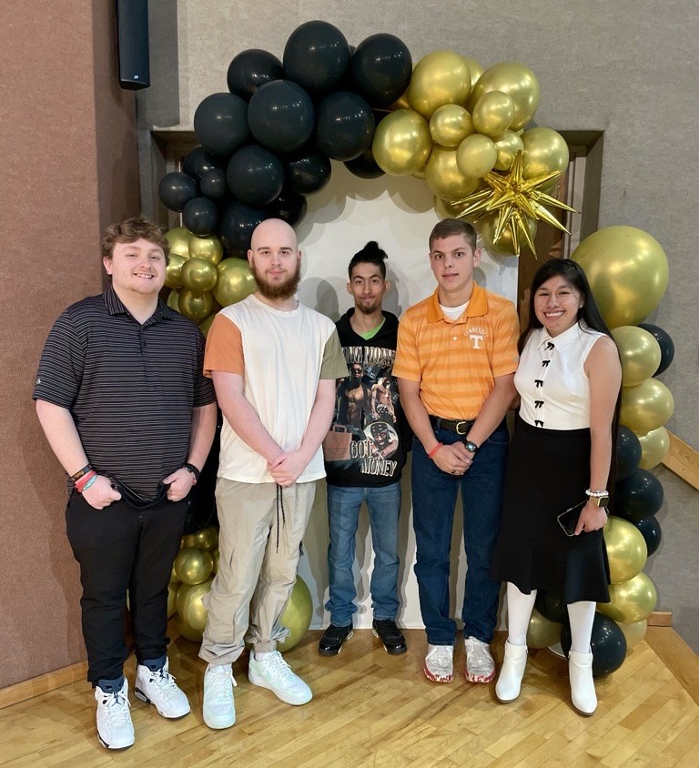Five people stand in front of a black and gold balloon arch, smiling at the camera during a Project SEARCH graduation. Four men and one woman, dressed in casual and semi-formal clothes, pose together on a wooden floor indoors.