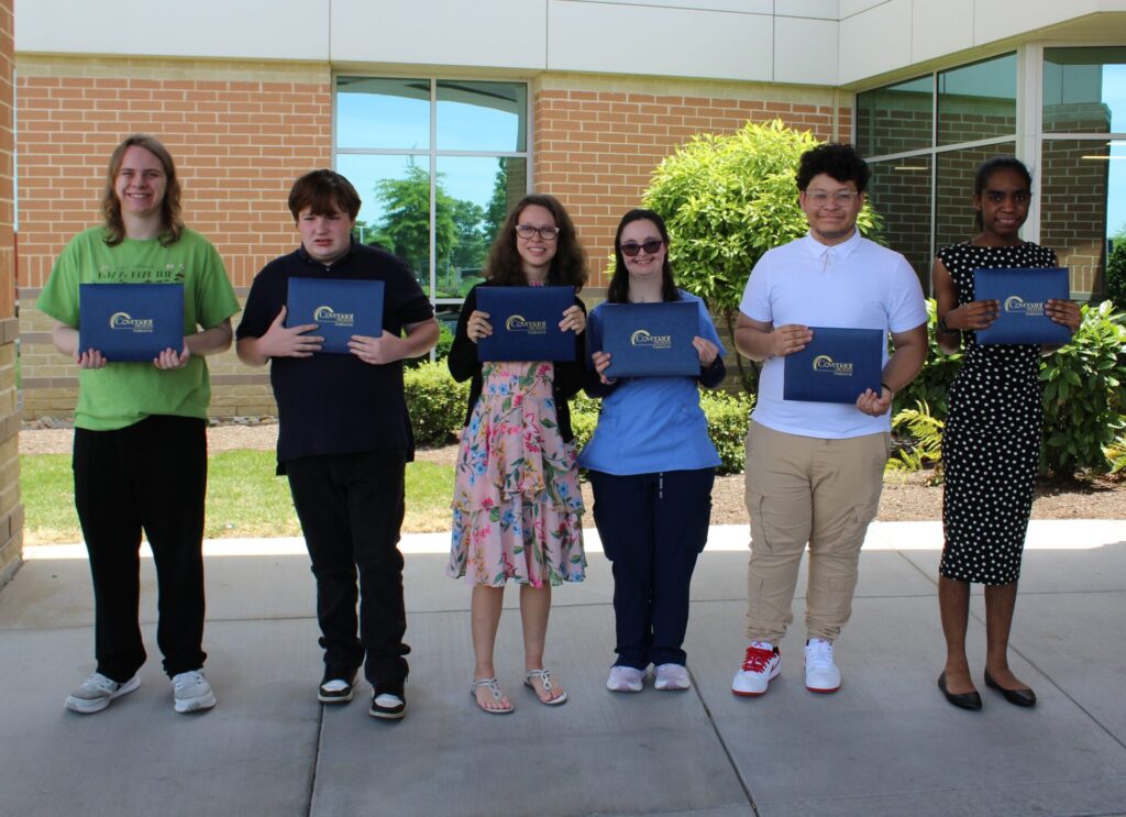 Six young adults stand in a row outside a building, smiling and holding certificates at their Project SEARCH graduation. Dressed in semi-formal attire, they appear to be celebrating this significant achievement together.