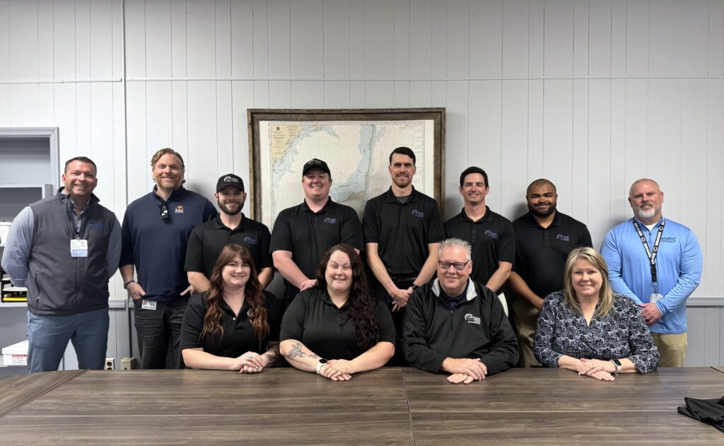 A group of eleven adults, eight men and three women, pose for a photo in a meeting room. Eight people stand in the back, three are seated at a table in front. Everyone is smiling. A map hangs on the wall behind them.