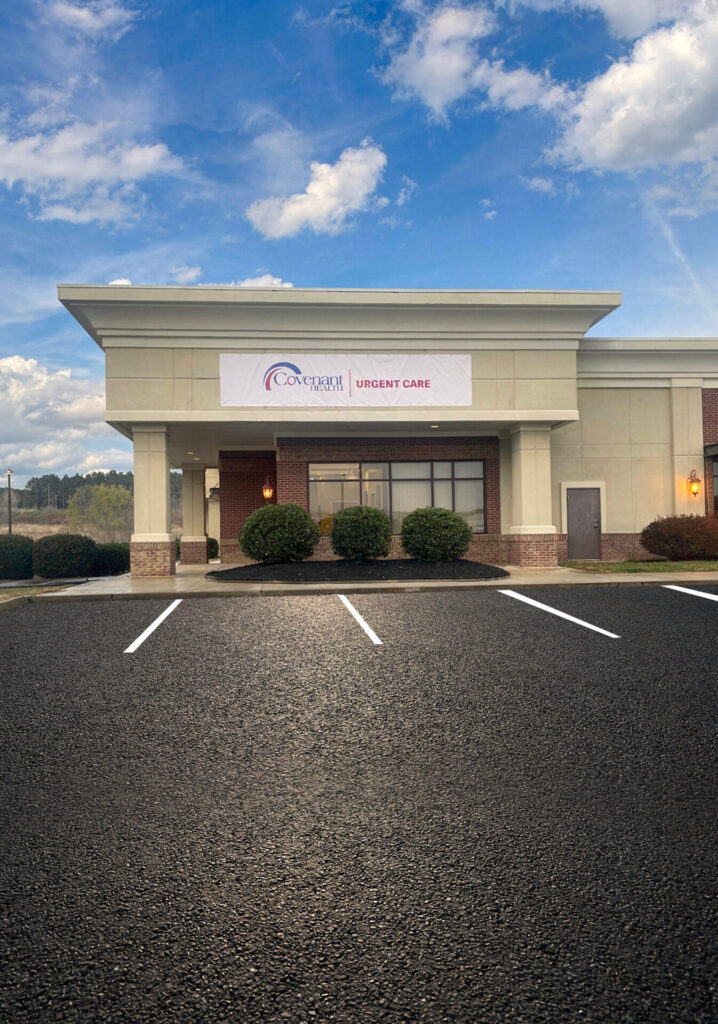 A small medical office building with a sign reading Urgent Care above the entrance, surrounded by bushes, and an empty parking lot in front, under a partly cloudy blue sky.