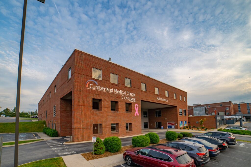 A brick building labeled Cumberland Medical Center with a safety grade rating, a pink ribbon symbol, and Main Entrance sign. Several cars are parked in front, and the sky is partly cloudy.
