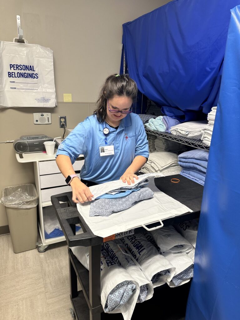 A woman wearing glasses and a blue uniform folds laundry on a black cart in a hospital.