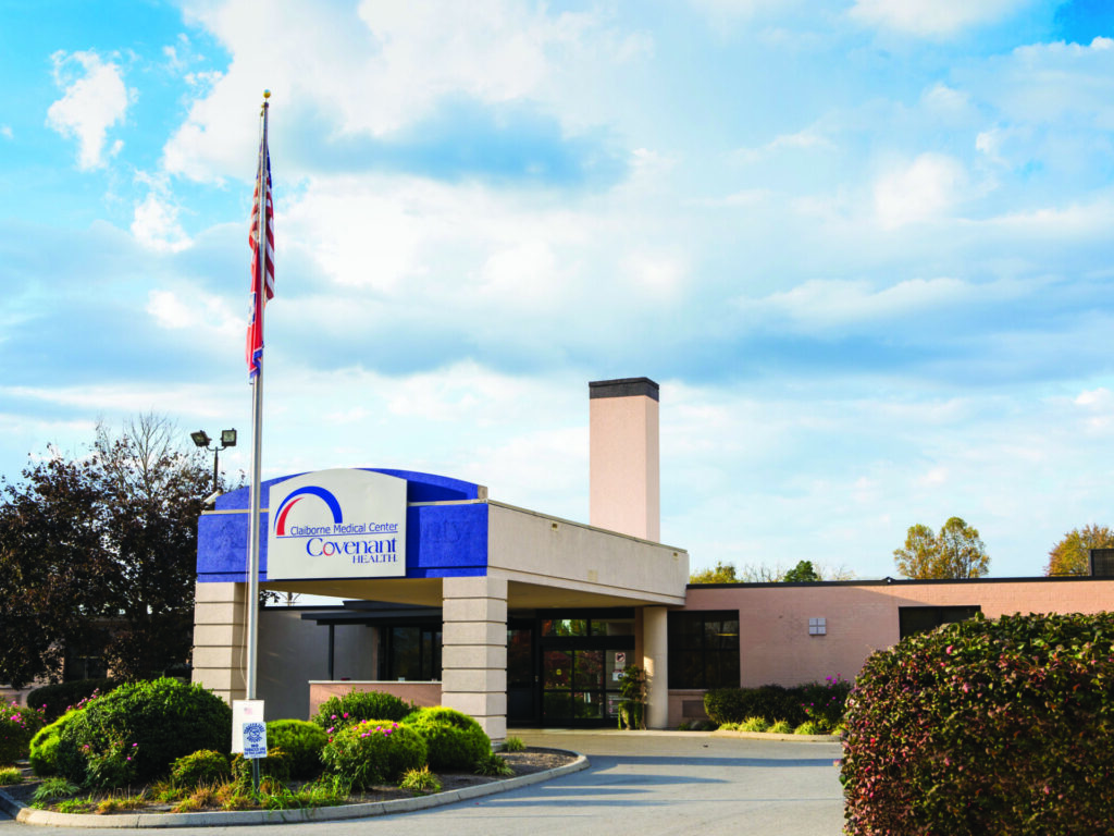 A medical center building with a blue and white sign reading Covenant Health near the entrance, surrounded by trimmed bushes and a U.S. flagpole, under a partly cloudy sky.