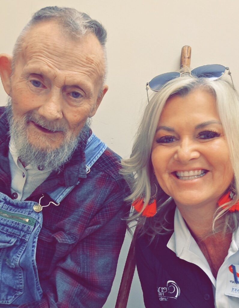 An older man with a gray beard and plaid shirt sits next to a smiling woman with blonde hair, red earrings, and sunglasses on her head. They are posed closely together indoors, reflecting the warmth of connected care management.
