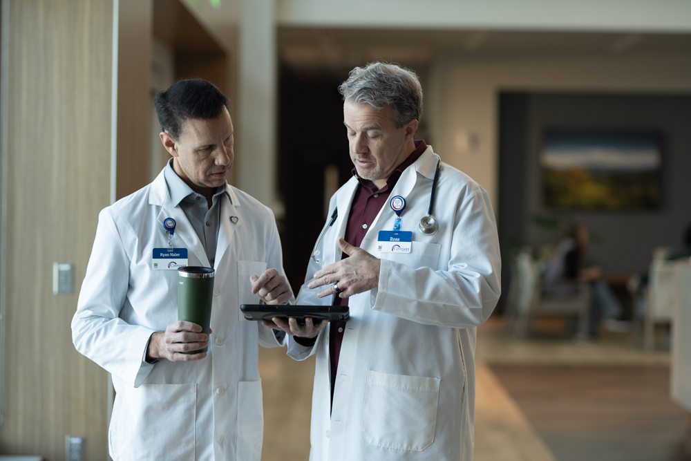 Two male doctors in white coats stand indoors, discussing information on a digital tablet. One holds a coffee tumbler, while the other gestures at the tablet. They appear focused and engaged in conversation.