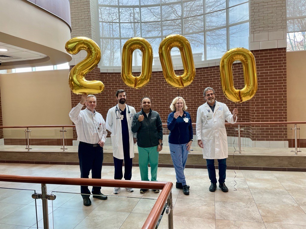 Five medical professionals stand indoors holding large gold balloons spelling &ldquo;2000,&rdquo; celebrating a milestone. The background features large windows, brick, and neutral-colored walls.