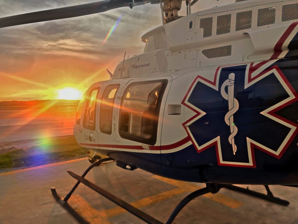 A medical helicopter with the Star of Life emblem is parked on a helipad at sunset, with sunlight reflecting off its windows and the sky glowing orange and yellow in the background.