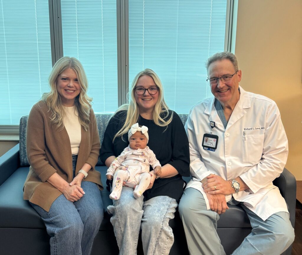 Three adults and a baby sit on a couch in an office or clinic. Two women, one holding the baby, sit next to a man in a white lab coat. All are smiling and facing the camera.