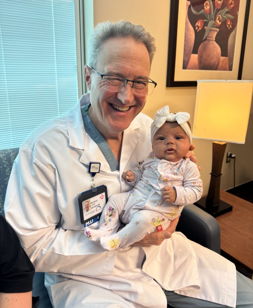 A smiling doctor in a white coat holds a baby dressed in a floral outfit and white headband, sitting together in a well-lit office with a table lamp and artwork in the background.